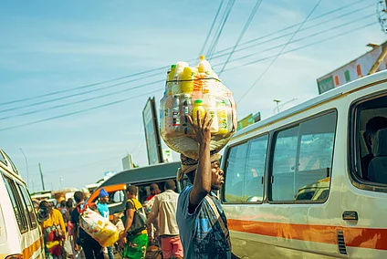 A bustling urban street scene with several white and orange minibuses parked and moving, likely serving as public transport. In the foreground, a person balances a large transparent container filled with colorful plastic bottles on their head. Other people walk or stand near the vehicles, suggesting a busy market or transport hub. Overhead, power lines and billboards are visible against a clear blue sky.