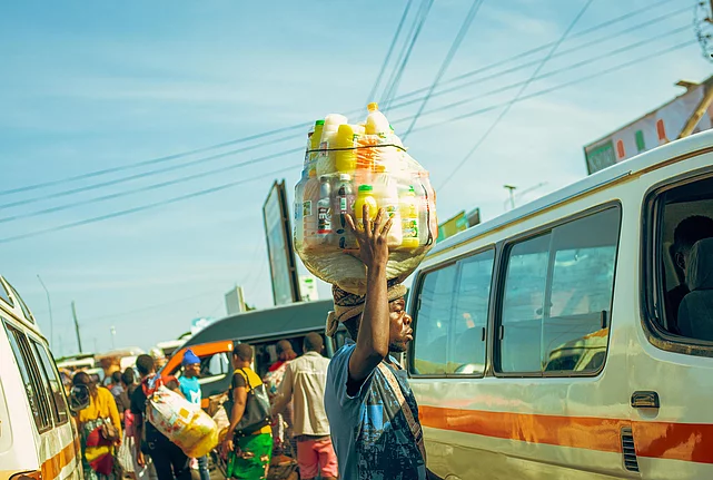 A bustling urban street scene with several white and orange minibuses parked and moving, likely serving as public transport. In the foreground, a person balances a large transparent container filled with colorful plastic bottles on their head. Other people walk or stand near the vehicles, suggesting a busy market or transport hub. Overhead, power lines and billboards are visible against a clear blue sky.