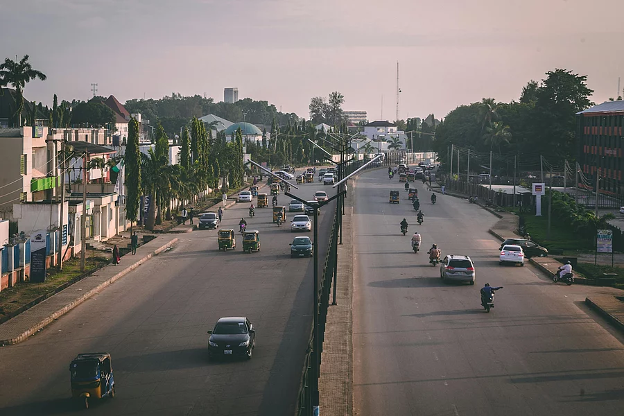A wide urban road with a central median, carrying cars, motorcycles, and auto rickshaws in both directions. On the left side, tall palm trees line the street beside low-rise buildings, while the right side features mixed-use commercial and residential structures, including a red and black building. Streetlights and scattered greenery frame the scene, with warm lighting suggesting early morning or late afternoon.