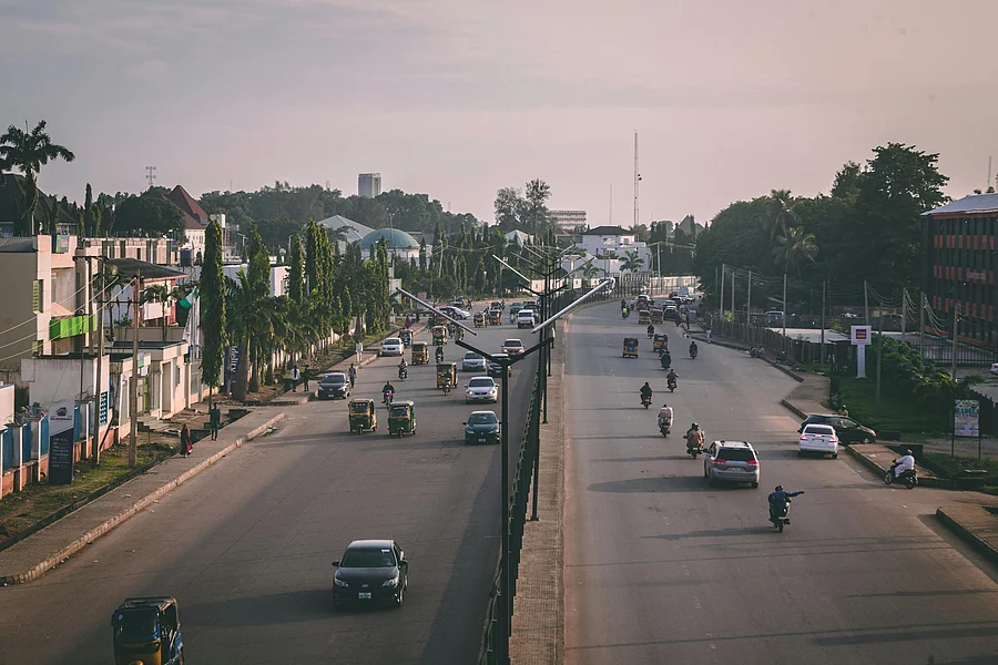 A wide urban road with a central median, carrying cars, motorcycles, and auto rickshaws in both directions. On the left side, tall palm trees line the street beside low-rise buildings, while the right side features mixed-use commercial and residential structures, including a red and black building. Streetlights and scattered greenery frame the scene, with warm lighting suggesting early morning or late afternoon.