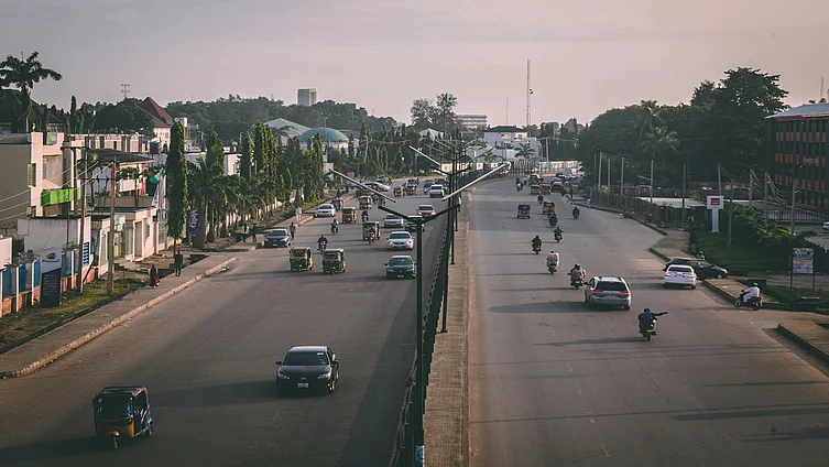 A wide urban road with a central median, carrying cars, motorcycles, and auto rickshaws in both directions. On the left side, tall palm trees line the street beside low-rise buildings, while the right side features mixed-use commercial and residential structures, including a red and black building. Streetlights and scattered greenery frame the scene, with warm lighting suggesting early morning or late afternoon.