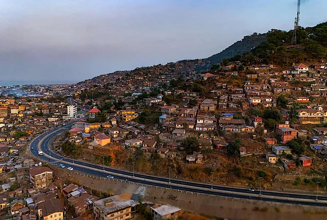 An aerial view of a densely populated hillside city with tightly packed houses in shades of brown, yellow, and red. A winding paved road cuts across the lower part of the scene. In the background, the city extends toward a coastal area with visible water and distant buildings. A tall communication tower stands on the hilltop, emitting a thin plume of smoke.