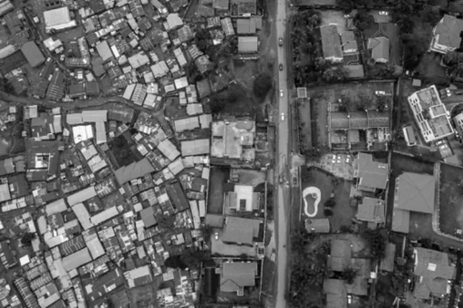 A black and white aerial photograph showing a stark contrast between two urban zones. On the left, a dense cluster of tightly packed buildings suggests an unplanned or informal settlement. On the right, more spacious plots with organized layouts and visible yards indicate a planned residential area. A central road divides the two zones, highlighting differences in land use, infrastructure, and urban planning
