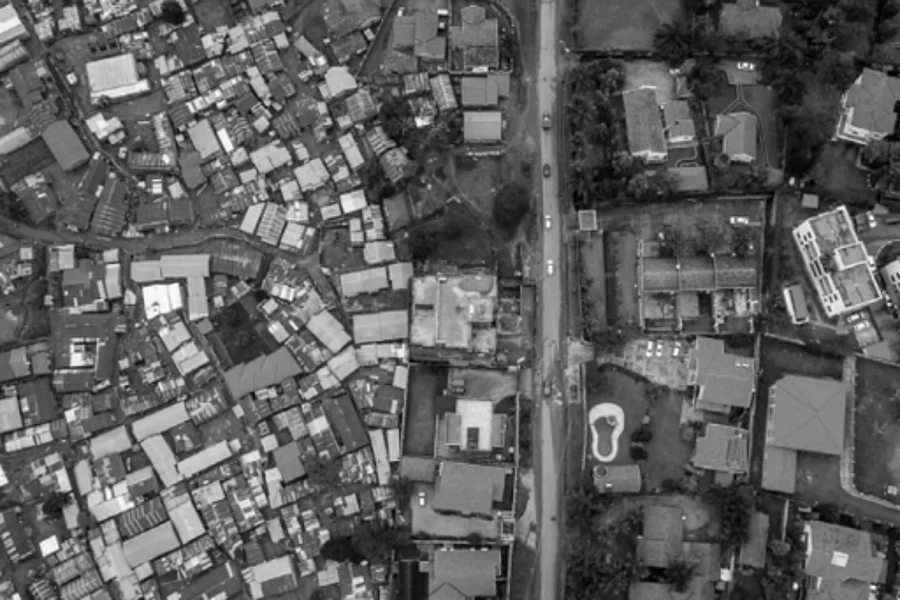 A black and white aerial photograph showing a stark contrast between two urban zones. On the left, a dense cluster of tightly packed buildings suggests an unplanned or informal settlement. On the right, more spacious plots with organized layouts and visible yards indicate a planned residential area. A central road divides the two zones, highlighting differences in land use, infrastructure, and urban planning