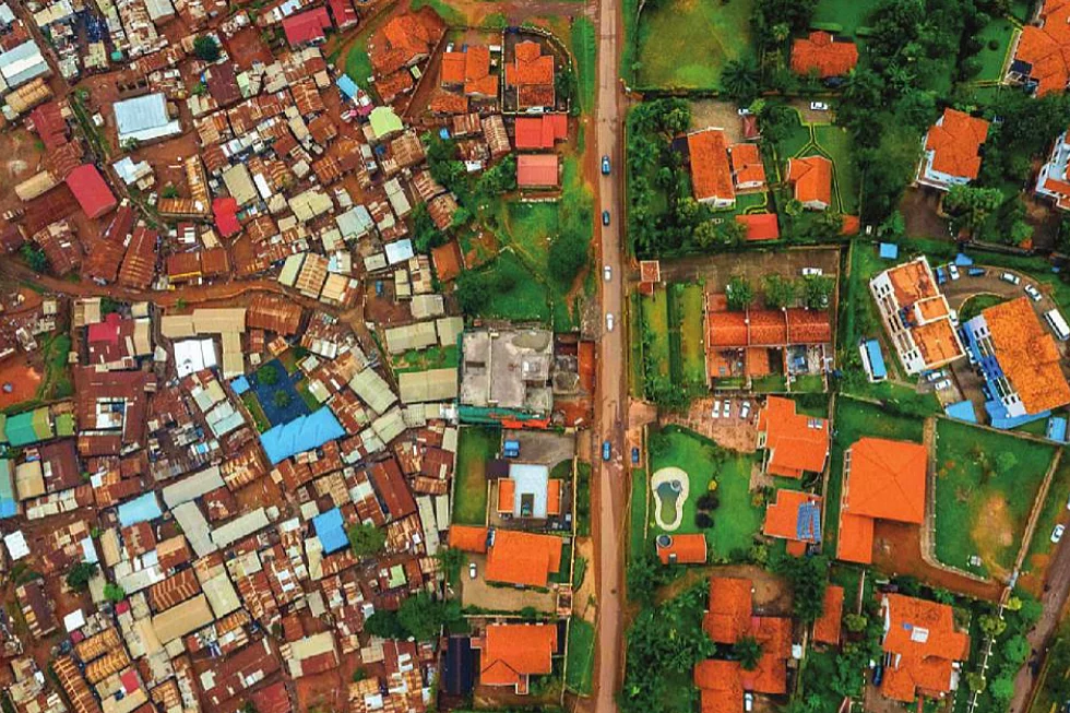 A black and white aerial photograph showing a stark contrast between two urban zones. On the left, a dense cluster of tightly packed buildings suggests an unplanned or informal settlement. On the right, more spacious plots with organized layouts and visible yards indicate a planned residential area. A central road divides the two zones, highlighting differences in land use, infrastructure, and urban planning