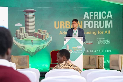 A speaker stands at a podium addressing attendees at the Africa Urban Forum 2019. Behind them, a large backdrop displays the text ‘Adequate Housing for All’ alongside a cityscape illustration with tall buildings and greenery. Rows of chairs and several audience members are visible in the foreground