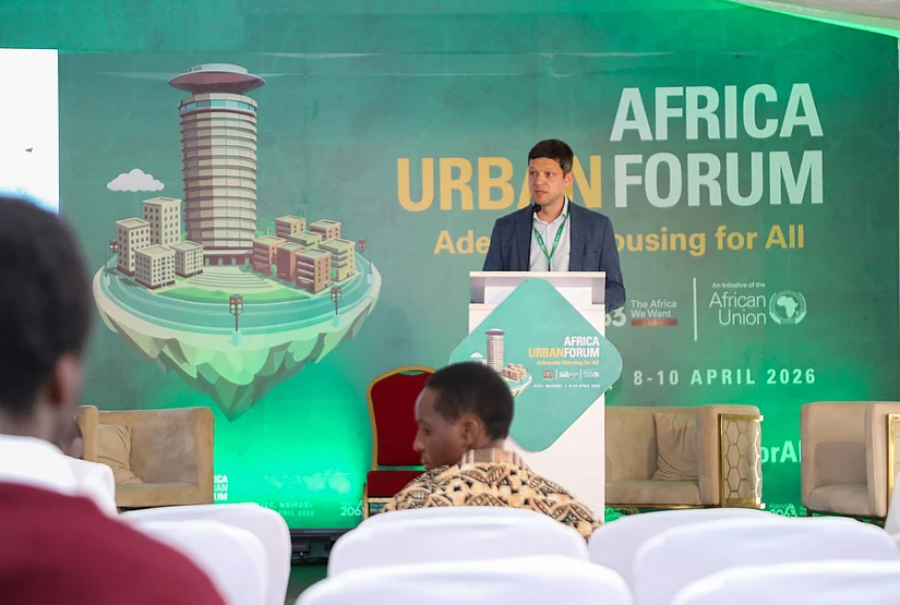 A speaker stands at a podium addressing attendees at the Africa Urban Forum 2019. Behind them, a large backdrop displays the text ‘Adequate Housing for All’ alongside a cityscape illustration with tall buildings and greenery. Rows of chairs and several audience members are visible in the foreground